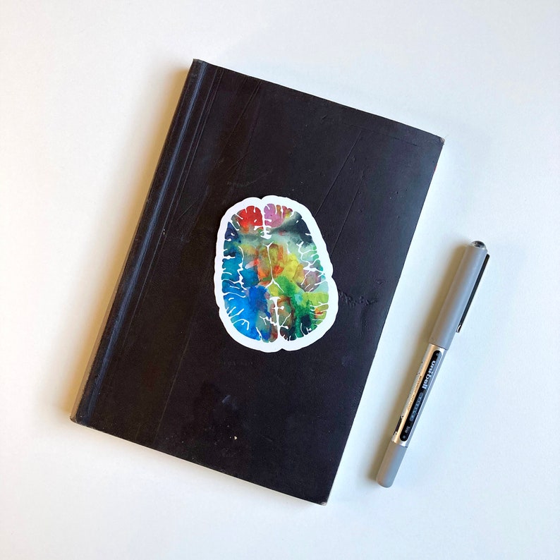 May include: A colourful watercolour brain sticker on a black notebook with a silver pen.