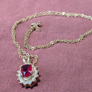 May include: Gold-toned necklace with a pendant featuring a large, oval, red gemstone surrounded by smaller, clear stones. The chain is delicate and rests on a mauve fabric surface. A classic jewelry piece.