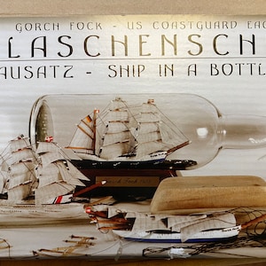 Puede incluir: Un kit de barco en una botella que presenta el Gorch Fock, un barco de vela alemán. El kit incluye una botella de vidrio transparente, una base de madera y un modelo detallado del barco. La caja presenta el texto "Gorch Fock - US Coastguard Eagle Flaschenschiff Bausatz - Ship in a Bottle Kit".