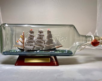 Op de afbeelding: Een gedetailleerd model van een zeilschip, de "Passat 1911", is ingesloten in een heldere glazen fles. Het schip heeft meerdere masten met witte zeilen en staat op een geschilderde blauwe basis. De fles staat op een rode houten standaard.