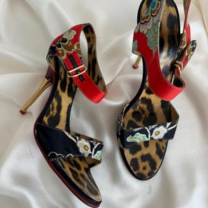 May include: A pair of high-heeled sandals with a leopard print and floral pattern. The sandals have a gold heel and a red strap.