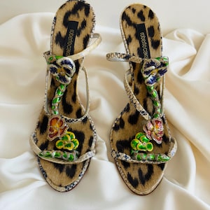 May include: A pair of high-heeled sandals with a leopard print design. The sandals have a floral embellishment with green, blue, and yellow flowers. The sandals are made by Roberto Cavalli.