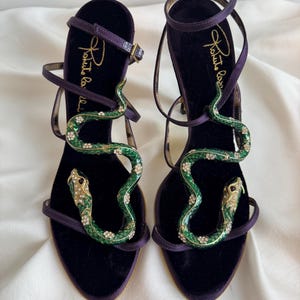 May include: A pair of purple velvet high-heeled sandals. The sandals feature a green snake design embellished with rhinestones. The straps are thin and wrap around the ankle. The brand name "Roberto Cavalli" is embroidered on the insole.