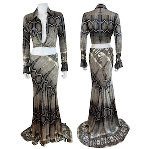May include: A two-piece outfit featuring a long-sleeve collared top with a front knot and a floor-length skirt. Both pieces display a brown, black, and cream snakeskin pattern. The top has long sleeves with flared cuffs.