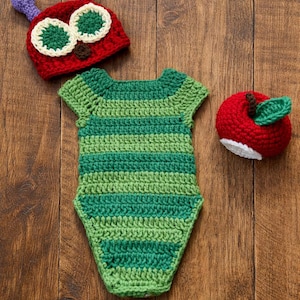 May include: A handmade crochet baby outfit set, featuring a green and white striped bodysuit, a red caterpillar hat with purple antennae, and a red apple with a green leaf. The set is designed for a baby.