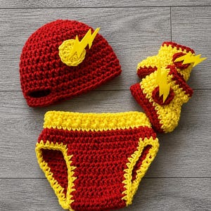 May include: A red and yellow crochet baby set, including a hat, diaper cover, and mittens. The hat and mittens feature a yellow lightning bolt design. The diaper cover has yellow trim.