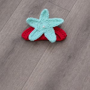 May include: A handmade crochet headband with a turquoise starfish and pearl centre, set on a red band. This accessory is ideal for babies and young children, adding a touch of whimsy to any outfit.