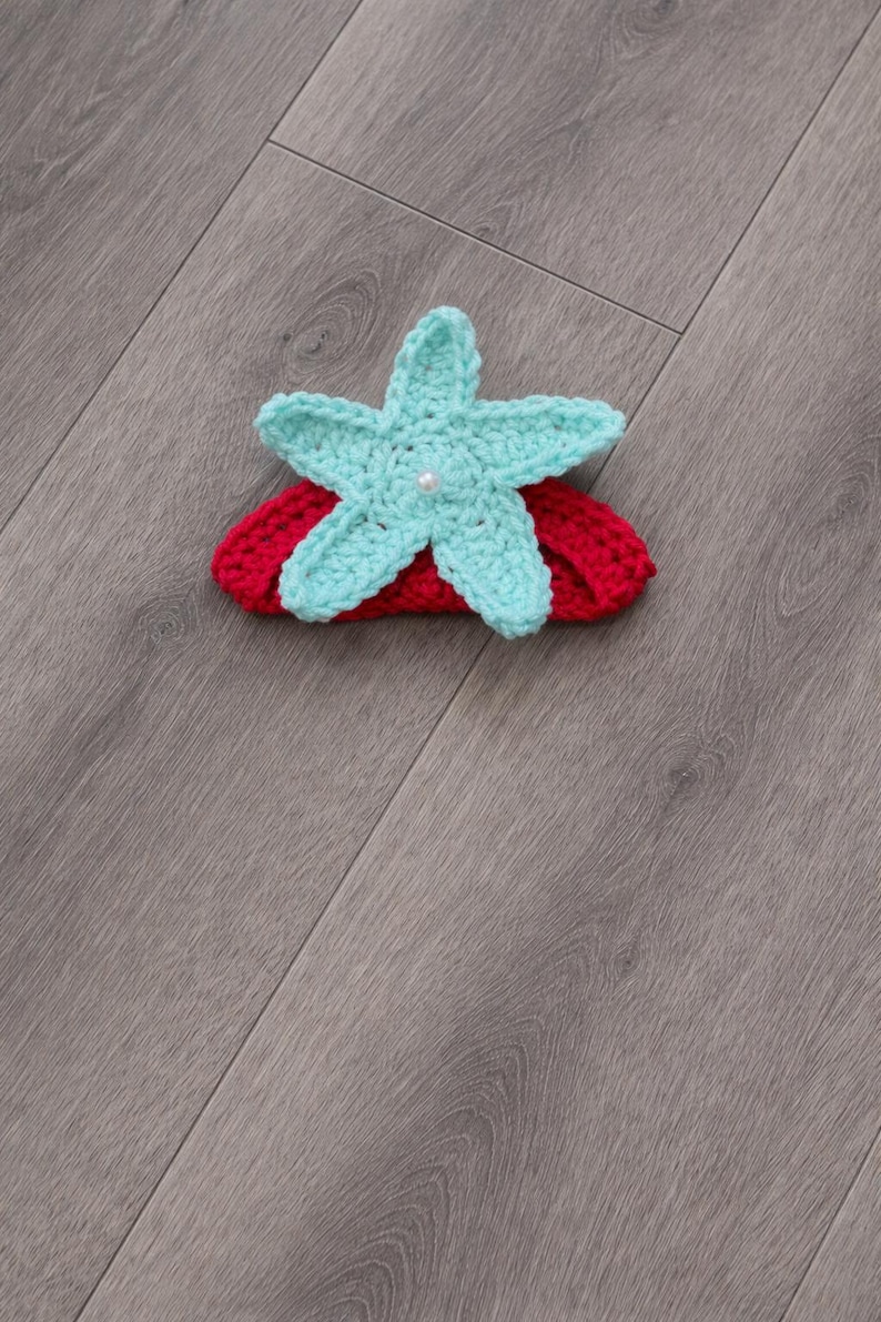 May include: A handmade crochet headband with a turquoise starfish and pearl centre, set on a red band. This accessory is ideal for babies and young children, adding a touch of whimsy to any outfit.