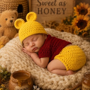 May include: A sleeping baby wearing a yellow crochet hat with ears, a red crochet shirt, and yellow crochet shorts. The baby is lying on a cream-colored knitted blanket. A wooden sign reads "Sweet as HONEY".