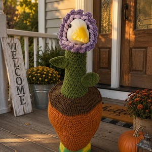 May include: A whimsical, handmade crochet duck flower pot cover. The duck has a white head, yellow beak, and a purple flower crown. The pot cover is orange, brown, and green, with a yellow and green base. A wooden sign that says "WELCOME" is in the background.
