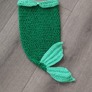 May include: A handmade, green crochet mermaid tail with a light green tail fin. The top edge has a scalloped trim and a small white bead. The tail is designed for a baby or toddler.
