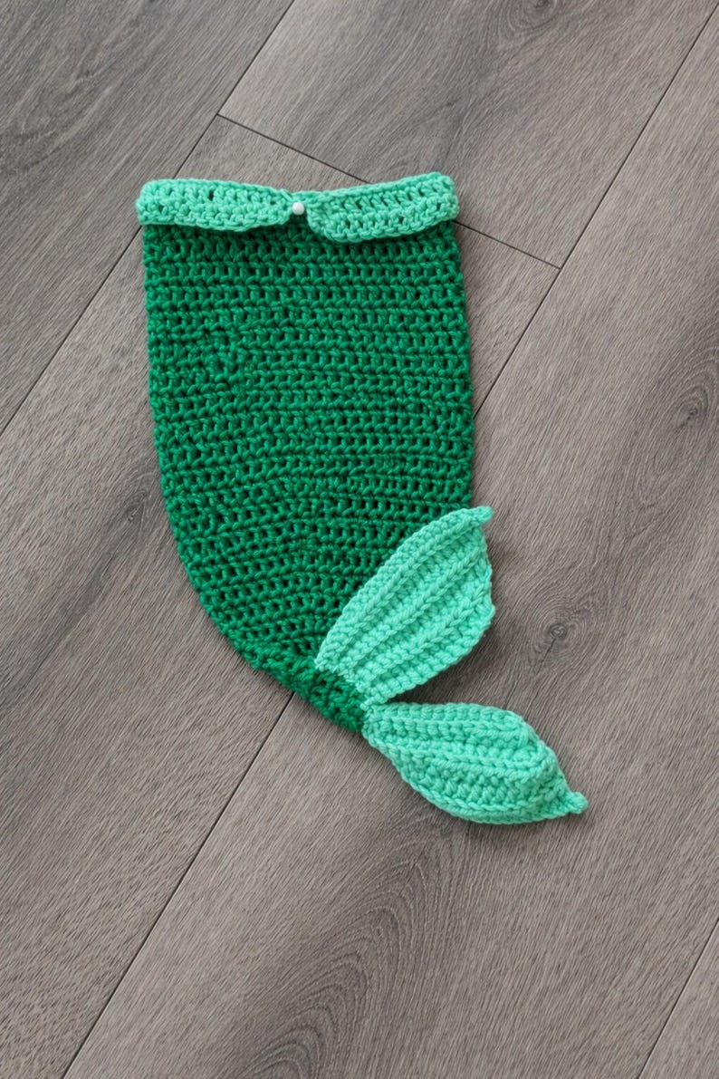 May include: A handmade, green crochet mermaid tail with a light green tail fin. The top edge has a scalloped trim and a small white bead. The tail is designed for a baby or toddler.