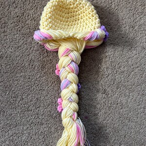 May include: A yellow crocheted hat with a long braided ponytail. The braid is made with yellow, pink, and purple yarn and has small crocheted flowers.