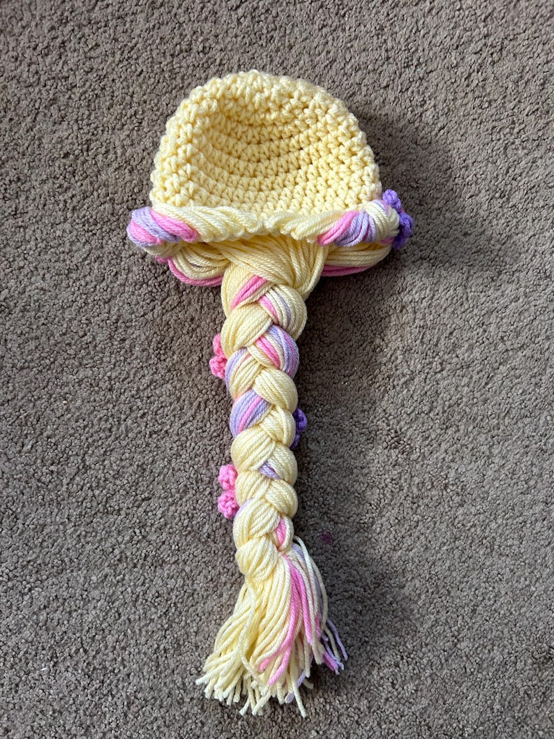May include: A yellow crocheted hat with a long braided ponytail. The braid is made with yellow, pink, and purple yarn and has small crocheted flowers.