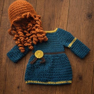 May include: A handmade crochet baby outfit in a teal blue color, featuring a dress with long sleeves and a matching hat with curly orange hair. The dress has yellow and brown accents, and is displayed on a wooden surface.