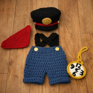 May include: A crocheted baby outfit in a train conductor theme. The set includes a black cap with a gold emblem, a red scarf, blue overalls with black suspenders, and a yellow pocket watch.
