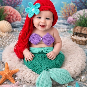 May include: A baby wearing a crochet mermaid costume, featuring a red wig with a blue flower, a purple top, and a green tail. The baby smiles, surrounded by starfish and pearls. The scene is set against a blue backdrop.
