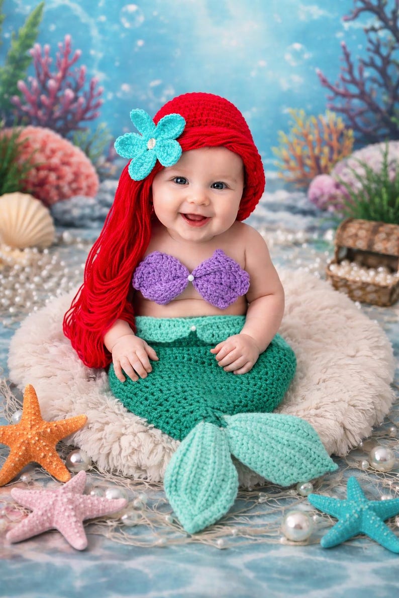 May include: A baby wearing a crochet mermaid costume, featuring a red wig with a blue flower, a purple top, and a green tail. The baby smiles, surrounded by starfish and pearls. The scene is set against a blue backdrop.