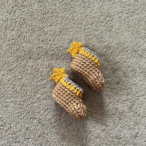 May include: A pair of brown crocheted baby booties with yellow star accents. The booties are on a light gray fuzzy surface.