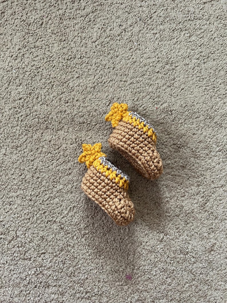 May include: A pair of brown crocheted baby booties with yellow star accents. The booties are on a light gray fuzzy surface.