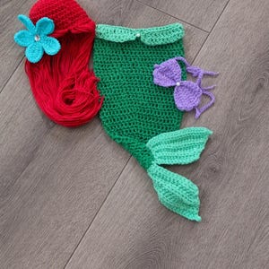 May include: A handmade crochet mermaid costume set in red, green, and purple. The set includes a red wig with a blue flower, a green top, a purple bikini top, and a green tail. Ideal for a baby's first photoshoot or a themed party.