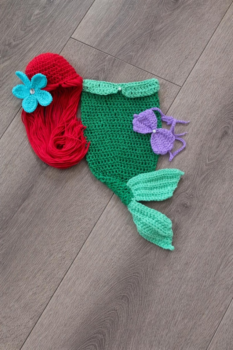 May include: A handmade crochet mermaid costume set in red, green, and purple. The set includes a red wig with a blue flower, a green top, a purple bikini top, and a green tail. Ideal for a baby's first photoshoot or a themed party.