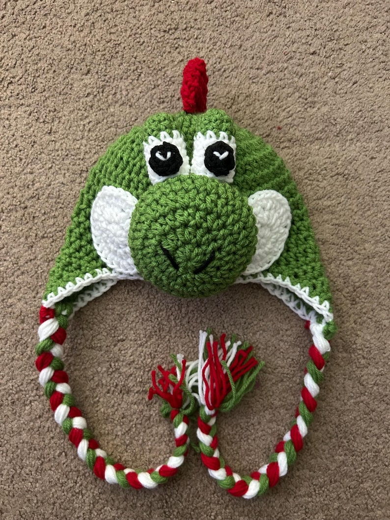 Puede incluir: Sombrero de crochet verde con una cara de dinosaurio, ojos blancos y una punta roja en la parte superior. El sombrero tiene un cord&oacute;n largo trenzado con hilo rojo, blanco y verde.
