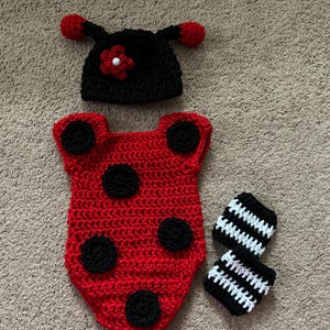 May include: A crocheted ladybug costume set for a baby, featuring a red and black bodysuit, a black hat with a red flower, and black and white striped leg warmers.