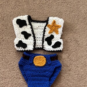 May include: Crocheted baby outfit inspired by a popular cowboy character. The outfit includes a white vest with black cow spots and a yellow star, and blue denim-style pants with a brown belt and a yellow button.
