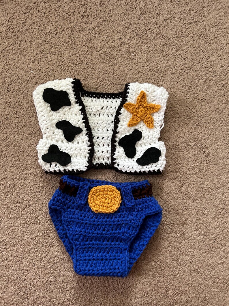 May include: Crocheted baby outfit inspired by a popular cowboy character. The outfit includes a white vest with black cow spots and a yellow star, and blue denim-style pants with a brown belt and a yellow button.