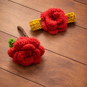 May include: Two handmade crochet items: a red rose with a green leaf and brown stem, and a red rose attached to a yellow headband. The items are displayed on a wooden surface.