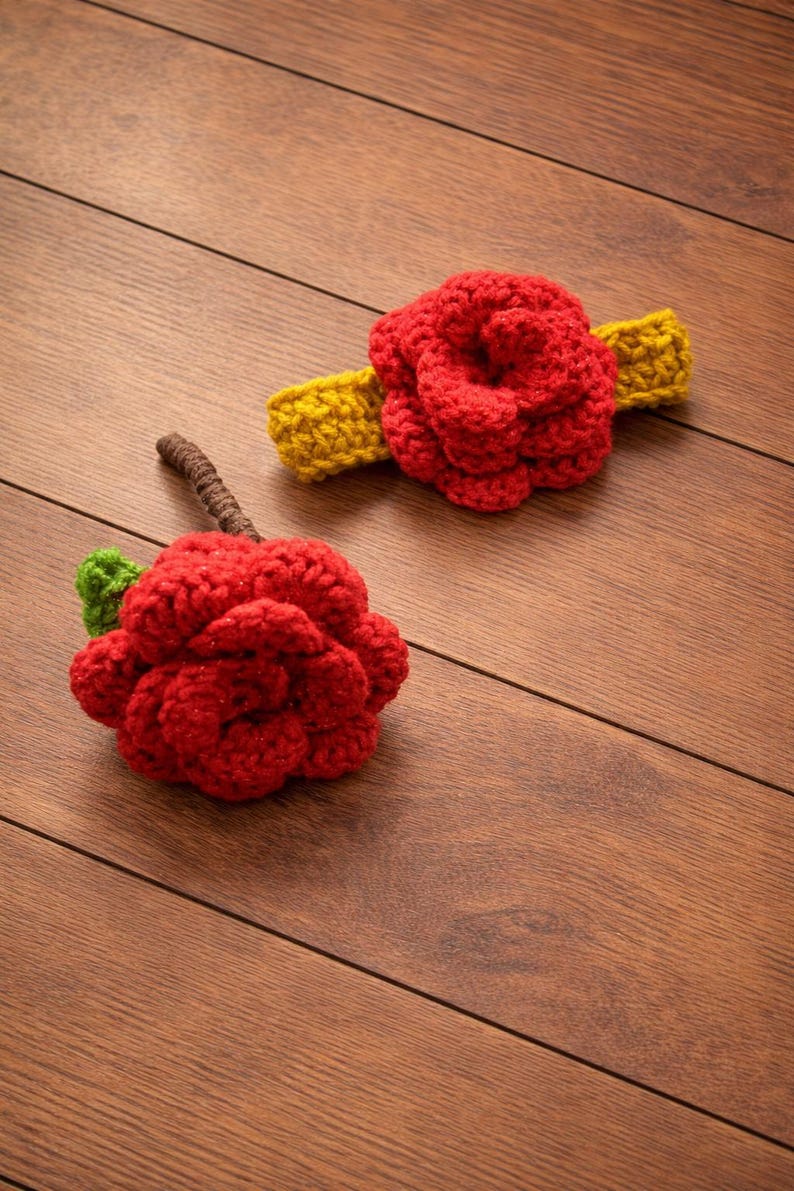 May include: Two handmade crochet items: a red rose with a green leaf and brown stem, and a red rose attached to a yellow headband. The items are displayed on a wooden surface.