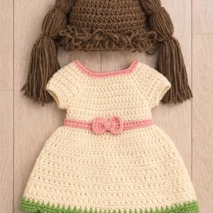 May include: A handmade crochet baby outfit, including a dress and a hat. The dress is cream-colored with pink trim and a bow, and a green scalloped hem. The hat is brown with pink accents and yarn pigtails.