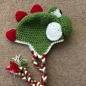Puede incluir: Sombrero de punto verde, rojo y blanco con forma de dinosaurio con un ojo blanco y un c&iacute;rculo blanco en el lateral. El sombrero tiene un cord&oacute;n largo trenzado.