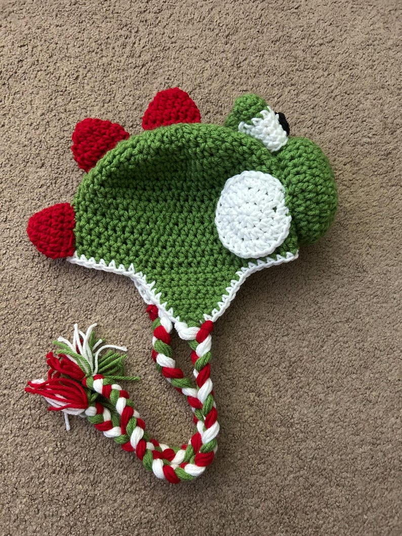 Puede incluir: Sombrero de punto verde, rojo y blanco con forma de dinosaurio con un ojo blanco y un c&iacute;rculo blanco en el lateral. El sombrero tiene un cord&oacute;n largo trenzado.