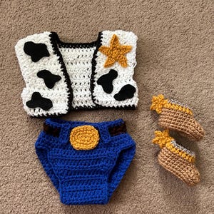 May include: A crocheted baby outfit inspired by a popular cowboy character. The set includes a white vest with black cow spots and a yellow star, blue denim diaper covers with a yellow sheriff's badge, and a pair of brown booties with yellow accents.