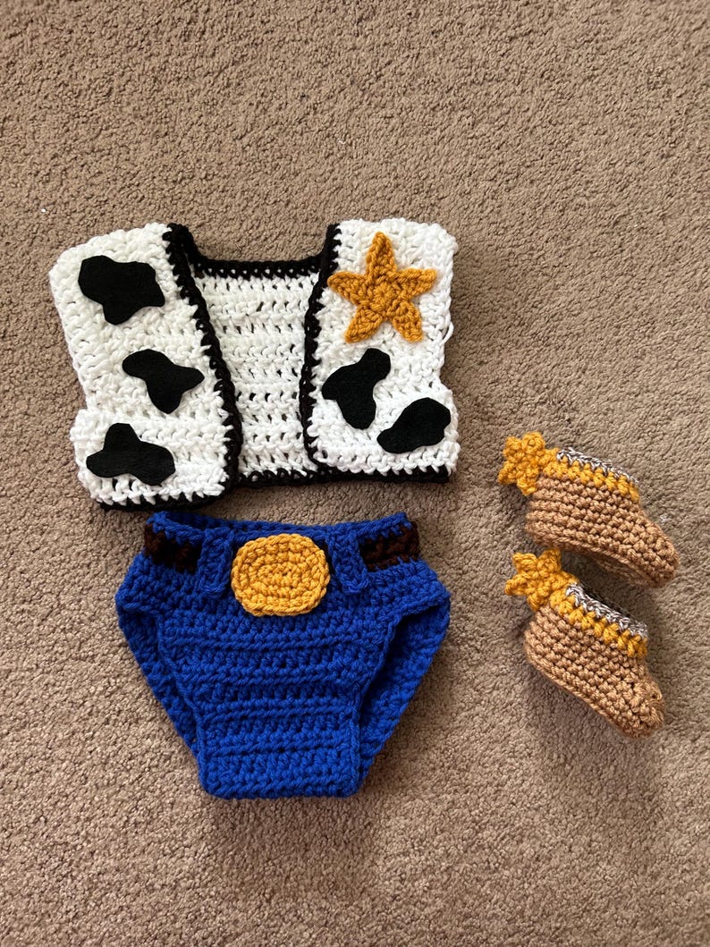 May include: A crocheted baby outfit inspired by a popular cowboy character. The set includes a white vest with black cow spots and a yellow star, blue denim diaper covers with a yellow sheriff's badge, and a pair of brown booties with yellow accents.