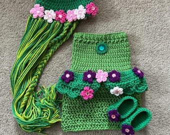 May include: A green crocheted baby outfit with a matching hat, shoes, and a long green yarn tail. The outfit is decorated with pink and purple flowers.