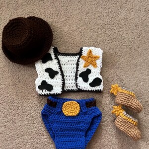 May include: A crocheted cowboy costume for a baby, featuring a brown hat, a white vest with black cow spots and a yellow star, blue denim diaper cover with a yellow buckle, and brown booties with yellow stars.