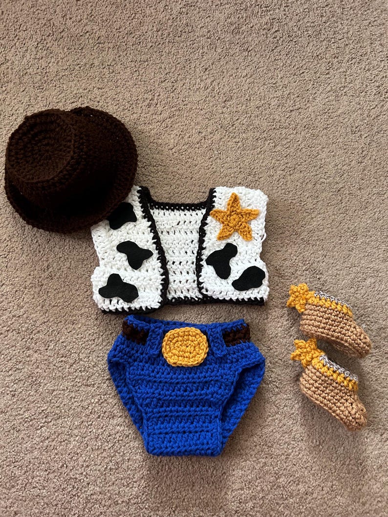 May include: A crocheted cowboy costume for a baby, featuring a brown hat, a white vest with black cow spots and a yellow star, blue denim diaper cover with a yellow buckle, and brown booties with yellow stars.