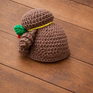 May include: A handmade brown crochet doll with a round head and body. A yellow string is wrapped around the neck. A green and red accent is attached to the side. The doll is lying on a wooden surface.
