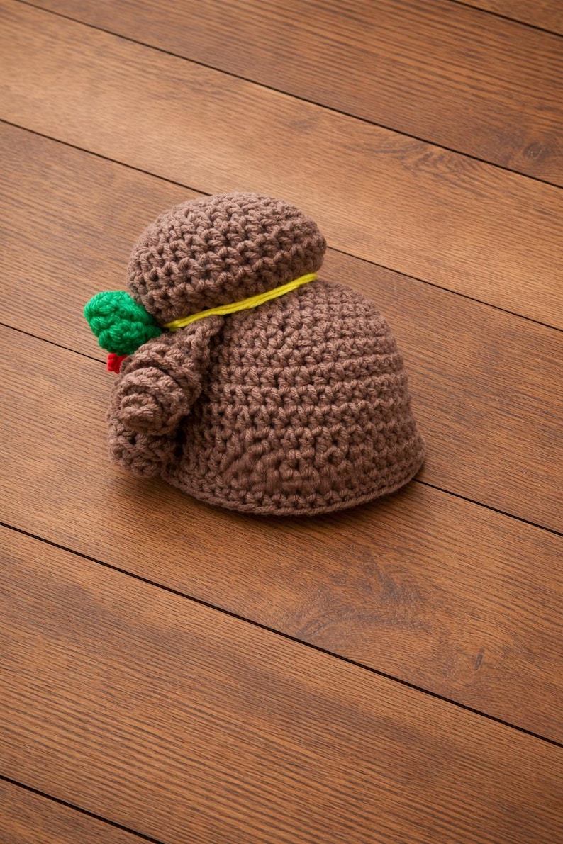 May include: A handmade brown crochet doll with a round head and body. A yellow string is wrapped around the neck. A green and red accent is attached to the side. The doll is lying on a wooden surface.
