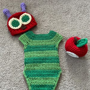 May include: A crocheted baby costume set featuring a green and white striped caterpillar bodysuit, a red hat with green eyes and a purple antenna, and a red apple hat.