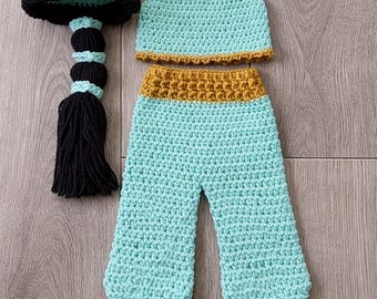 May include: Crocheted baby outfit in a teal color, featuring a top, pants, and a hat with a black braid. The top and pants have a gold-colored trim. The hat is black with teal and gold accents. A charming handmade baby costume.