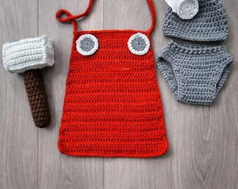 May include: A handmade crochet baby outfit in a Thor theme. The set includes a red tunic with gray and white accents, a gray hat with a white wing, gray diaper cover, and a crochet hammer. The outfit is displayed on a wooden surface.