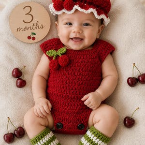 May include: A baby wearing a handmade red crochet outfit with a matching hat, adorned with green leaves and red cherries. The outfit includes a romper and leg warmers. A wooden disc with the text "3 months" and cherry illustrations is also present.