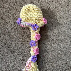 May include: A yellow crocheted hat with a long braided ponytail. The braid is decorated with pink and purple flowers.