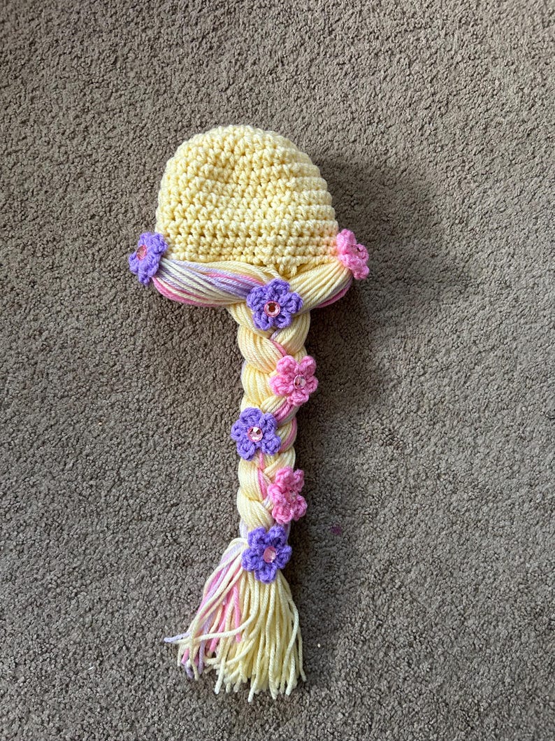May include: A yellow crocheted hat with a long braided ponytail. The braid is decorated with pink and purple flowers.