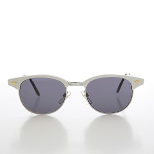 Puede incluir: Gafas de sol con montura plateada y lentes gris oscuro. Las monturas tienen un sutil acento dorado en las patillas.