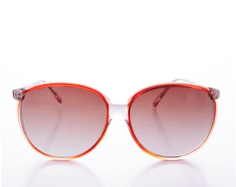 May include: Round sunglasses with a gradient brown tint. The frames are a translucent orange with clear arms. The lenses have a brown gradient. The sunglasses are set against a white background.